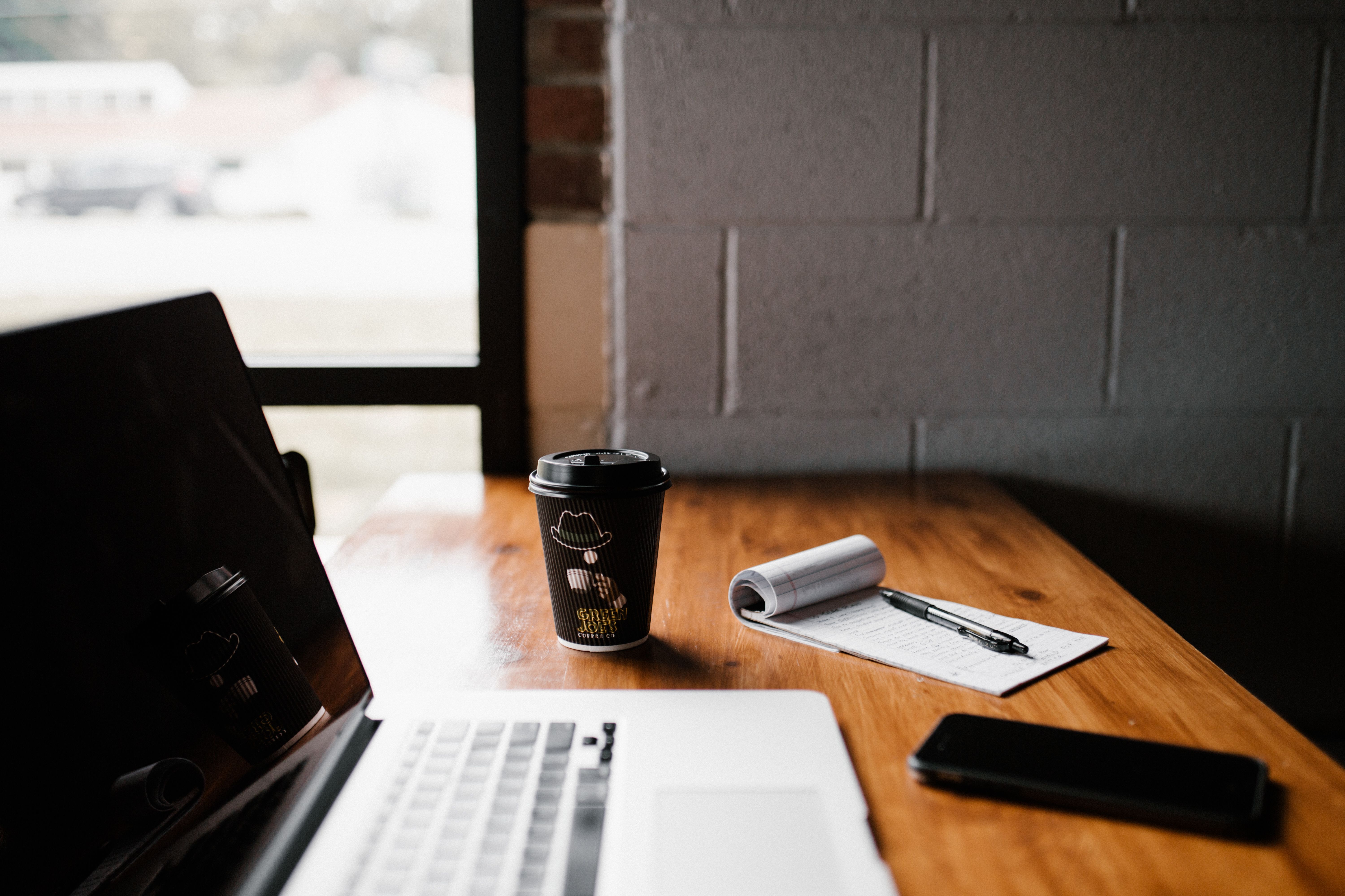 Laptop, notebook, and coffee on a desk beside a window